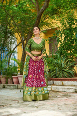 Woman in a green and pink traditional outfit standing outdoors with greenery around.