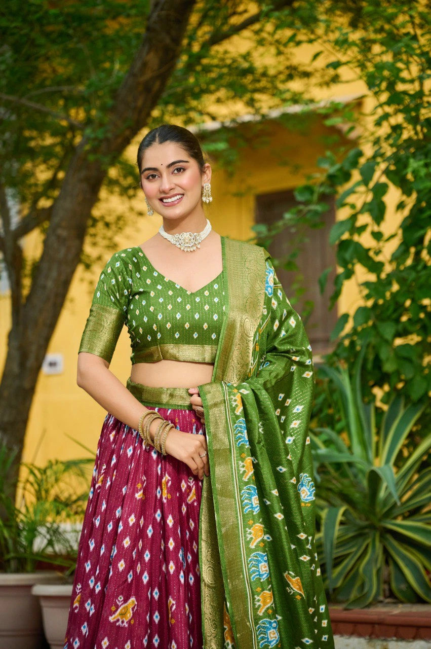 Woman in a green and pink traditional outfit standing outdoors with plants and a yellow wall in the background