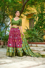 Woman in a green and pink traditional outfit standing outdoors with greenery around.