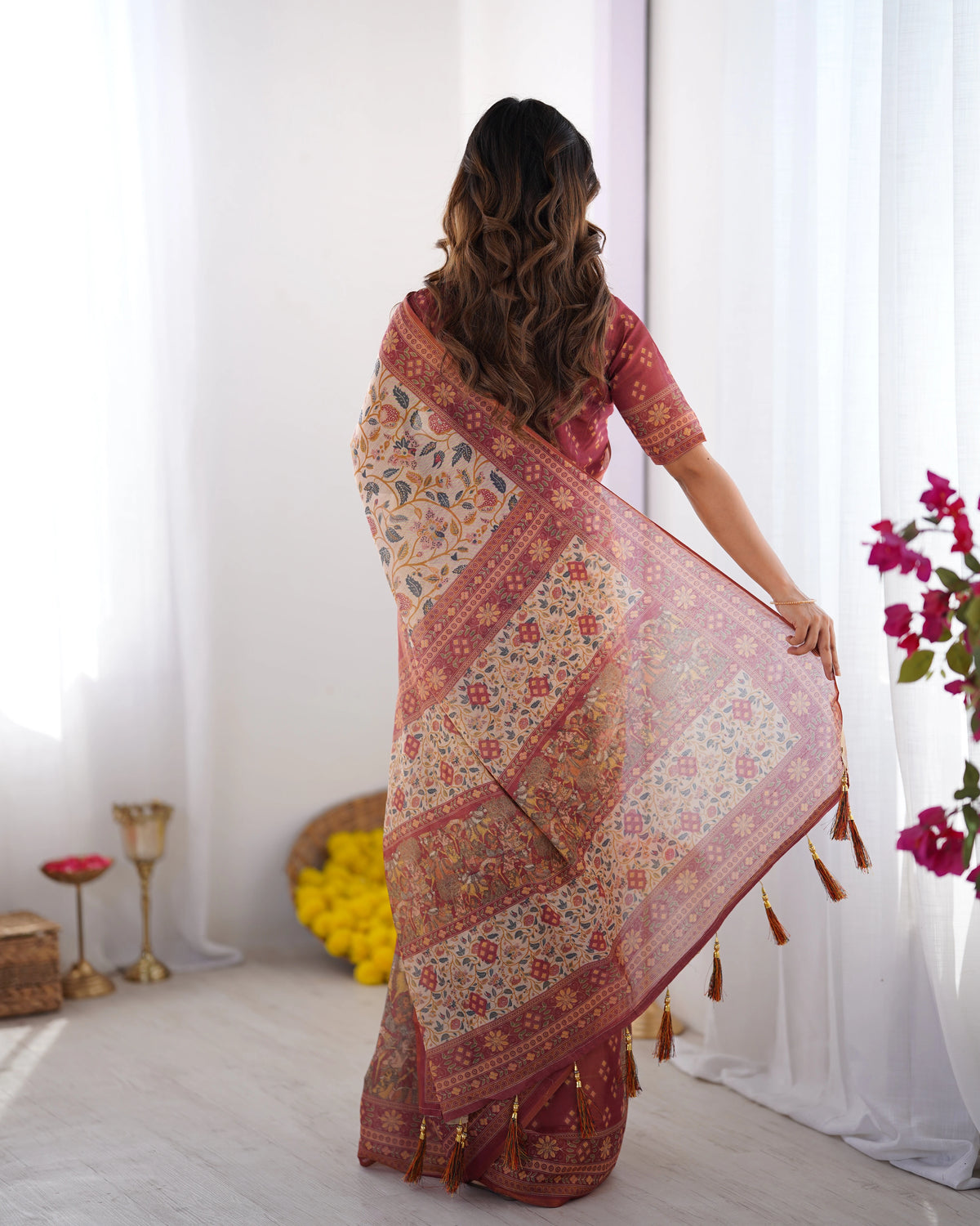 Woman wearing a floral saree in a room with white curtains and decorative items.
