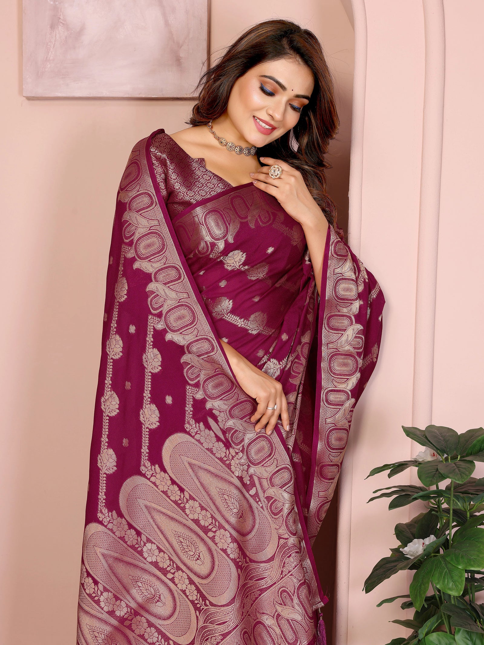 Woman wearing a maroon saree with intricate patterns, standing against a light pink wall.