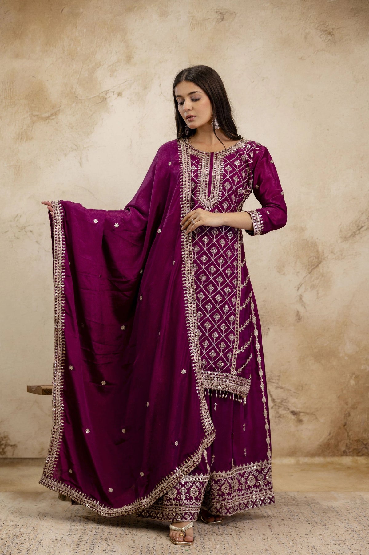 Woman wearing a purple traditional outfit with intricate designs against a beige wall.