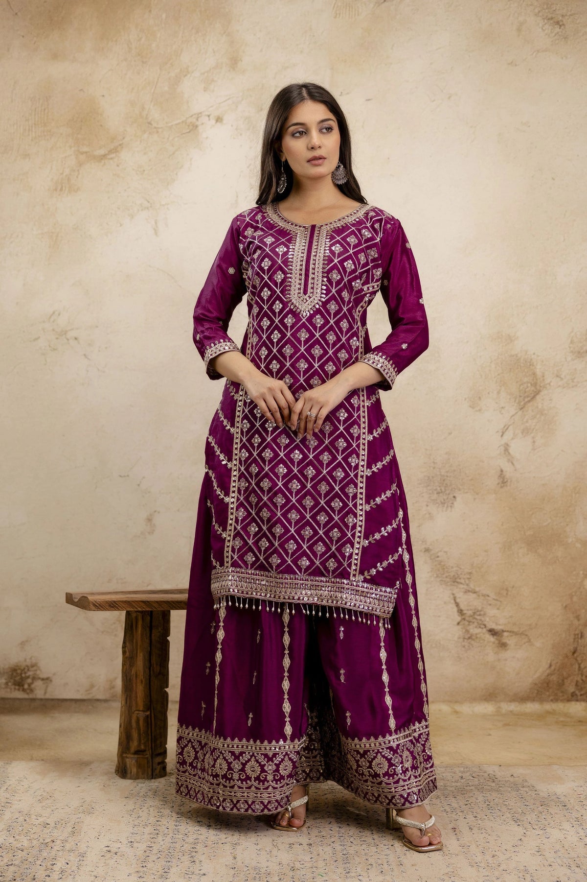 Woman wearing a purple traditional outfit with intricate patterns against a beige wall.