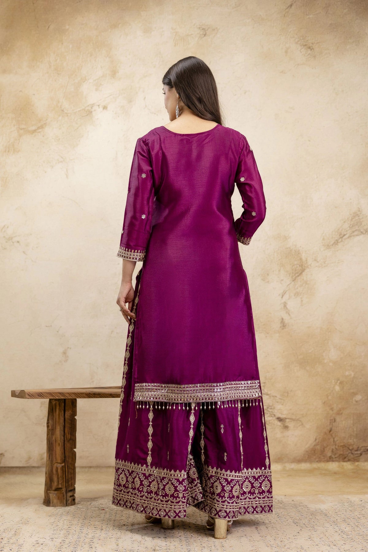 Woman wearing a purple traditional outfit with intricate patterns, standing against a beige wall.