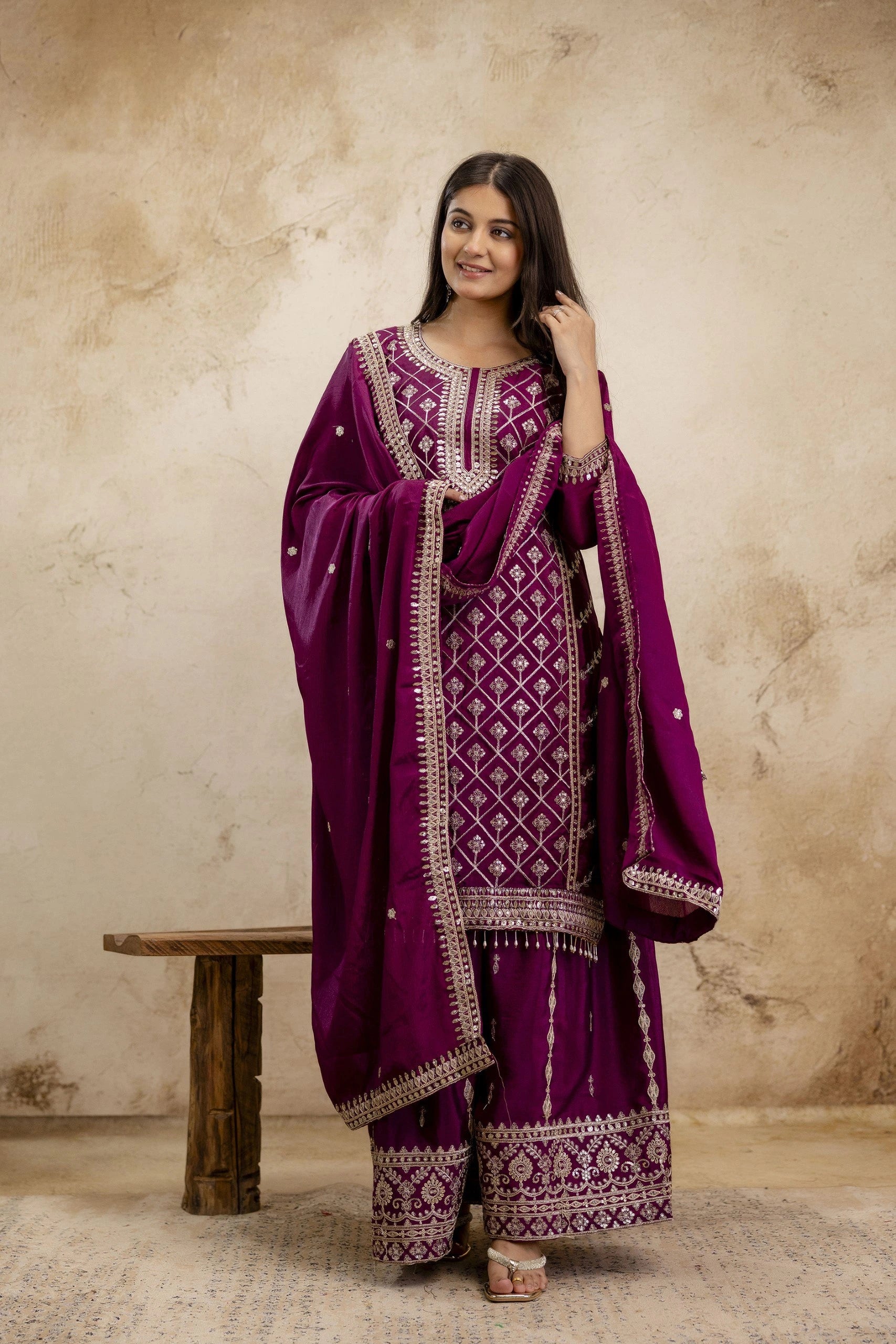 Woman wearing a traditional purple outfit with intricate patterns against a beige wall.