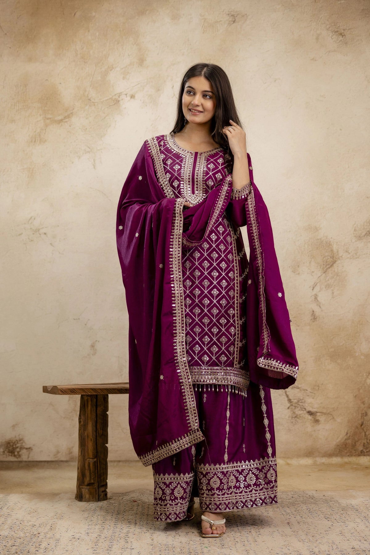 Woman wearing a traditional purple outfit with intricate patterns against a beige wall.