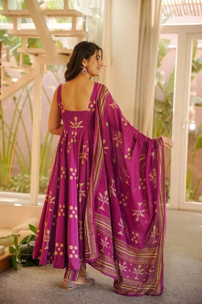 Woman in a purple traditional outfit with gold patterns standing in a room with large windows.