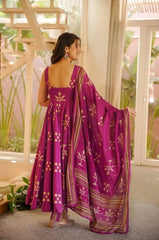 Woman in a purple traditional outfit with gold patterns standing in a room with large windows.