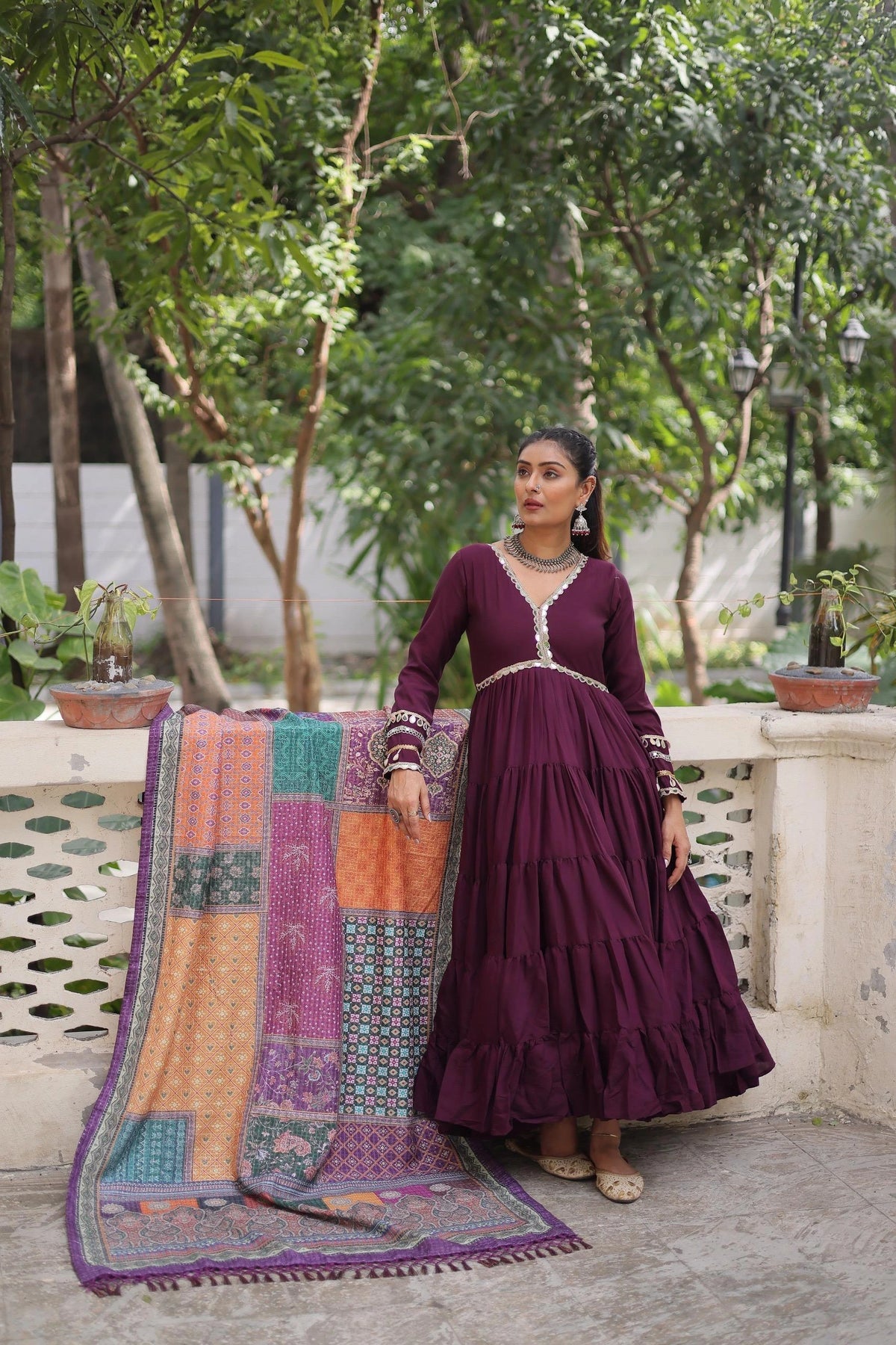 Woman in a long dark purple dress standing next to a colorful quilt on a stone ledge with greenery in the background.