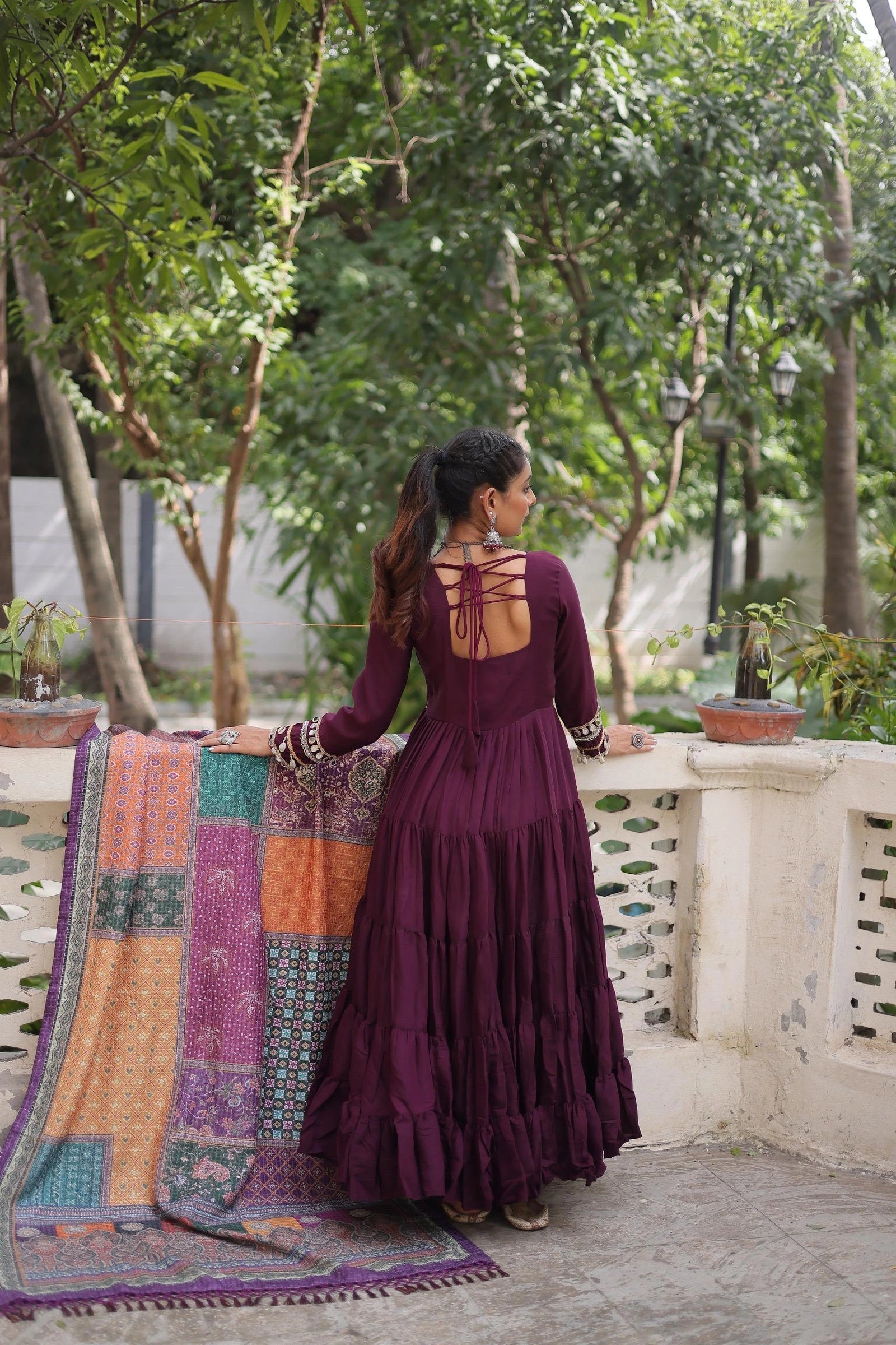 Woman in a purple dress standing on a balcony with a colorful quilt and greenery.
