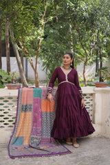 Woman in a long dark purple dress standing next to a colorful quilt on a stone ledge with greenery in the background.