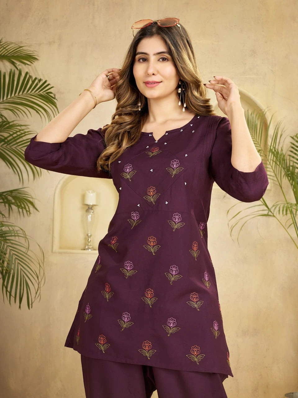 Woman wearing a purple embroidered outfit against a beige wall with green plants.