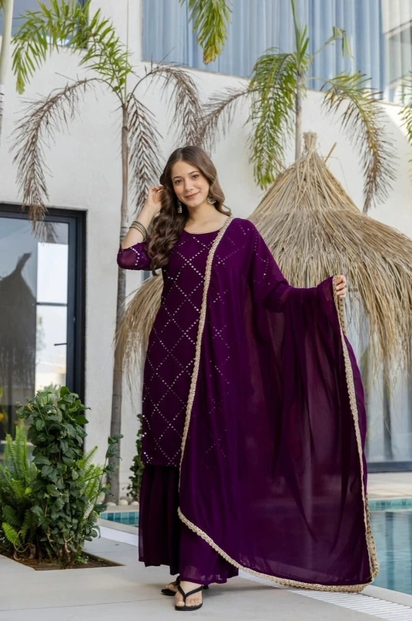 Woman in a purple traditional outfit standing outdoors with plants and a building in the background