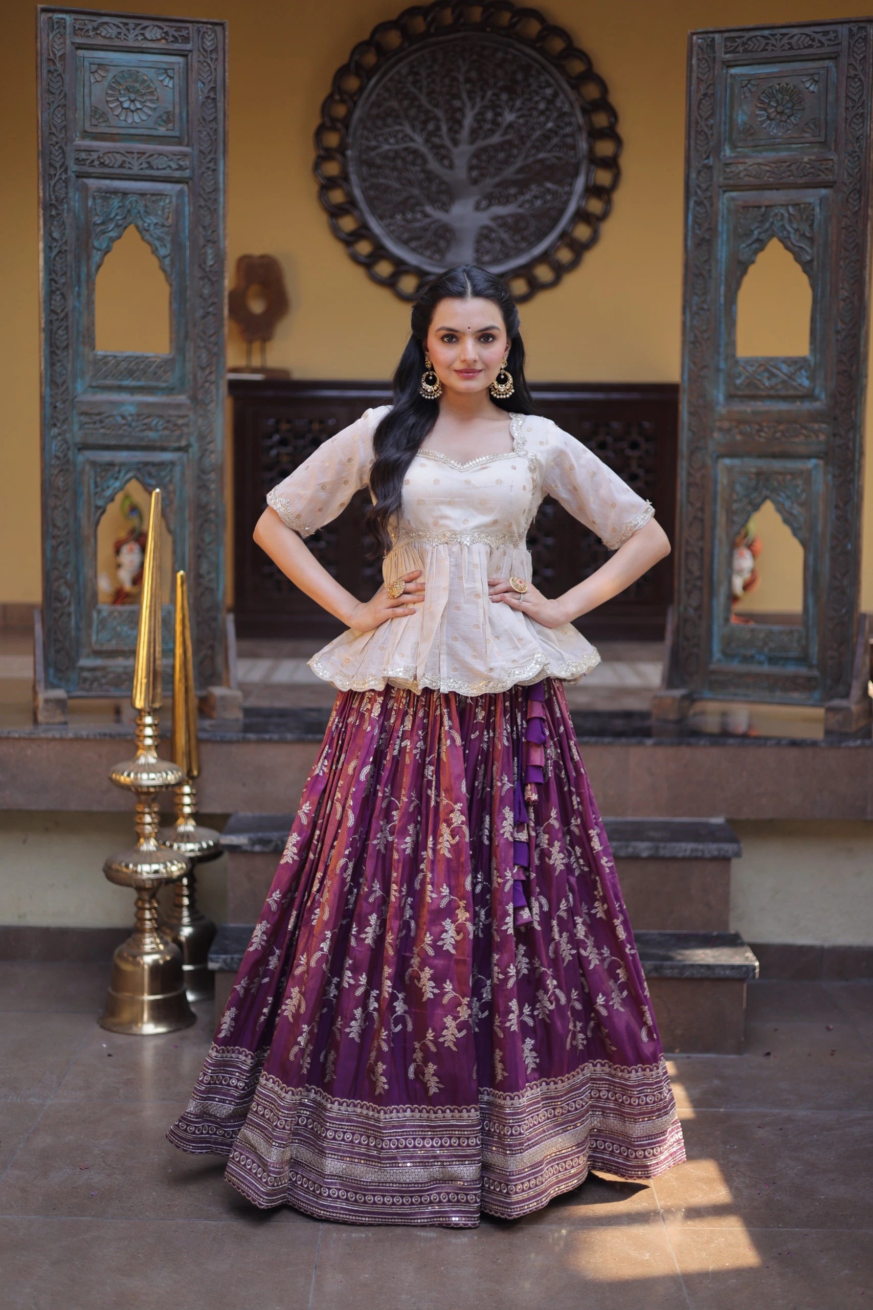 Woman in a traditional outfit with a white top and purple skirt standing in a decorated room.