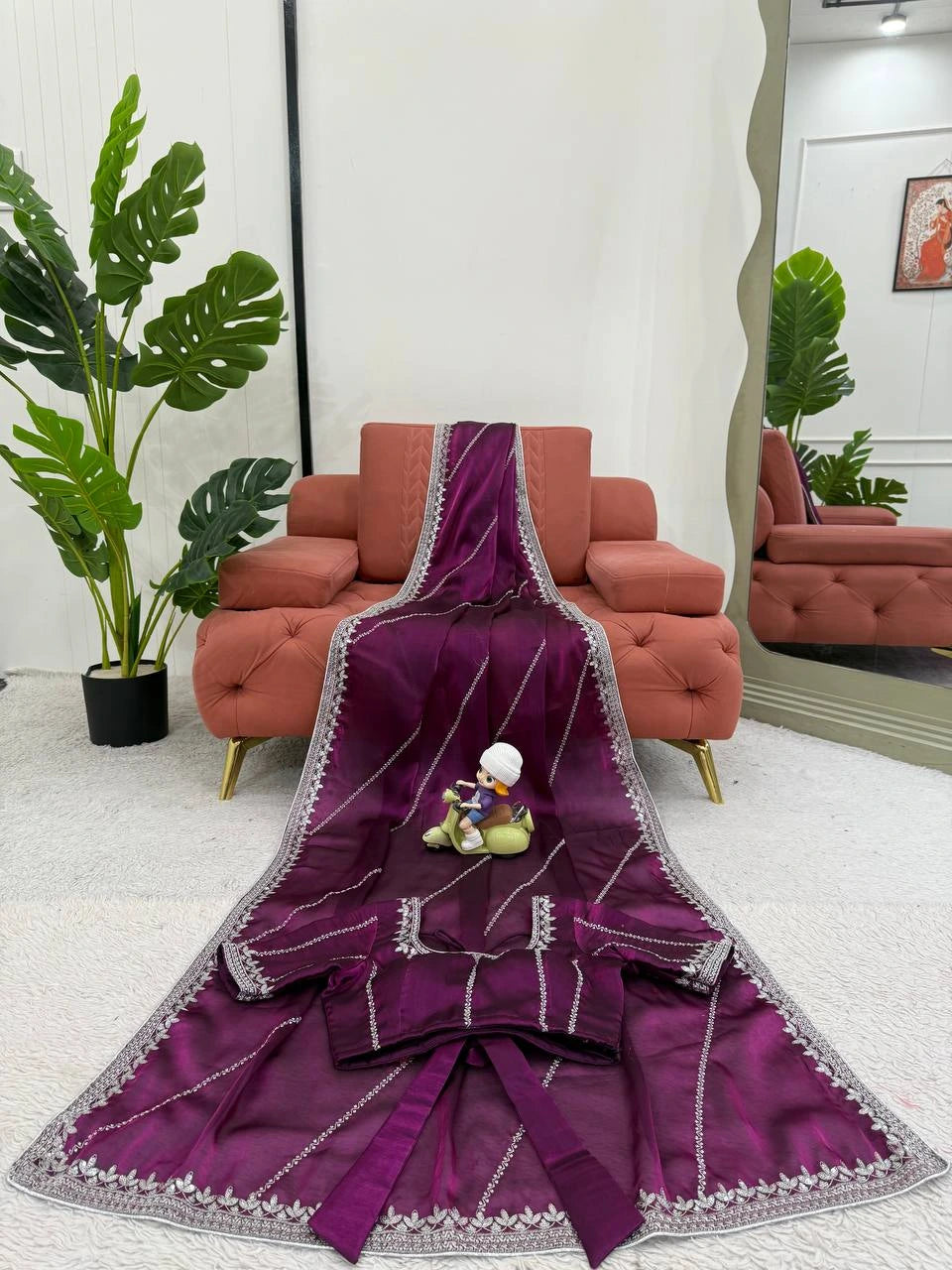 Purple embroidered fabric with a small figurine on a white surface, with a red sofa and green plants in the background.
