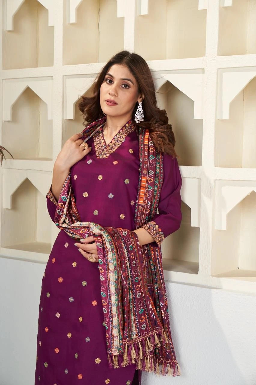 Woman in a purple traditional outfit with a colorful scarf against a decorative wall.