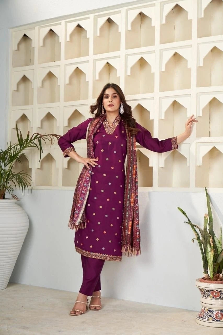 Woman in a purple traditional outfit with a patterned scarf standing against a decorative wall.