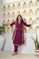 Woman in a purple traditional outfit with a patterned scarf standing against a decorative wall.