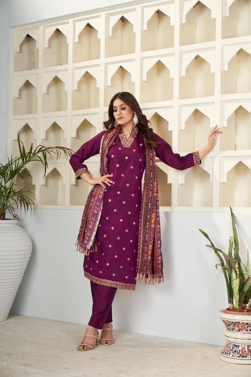 Woman in a purple traditional outfit with a patterned scarf standing against a decorative wall.