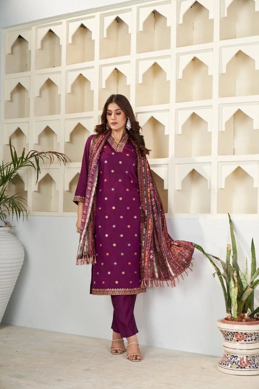 Woman in a purple traditional outfit with a patterned scarf standing against a decorative wall.