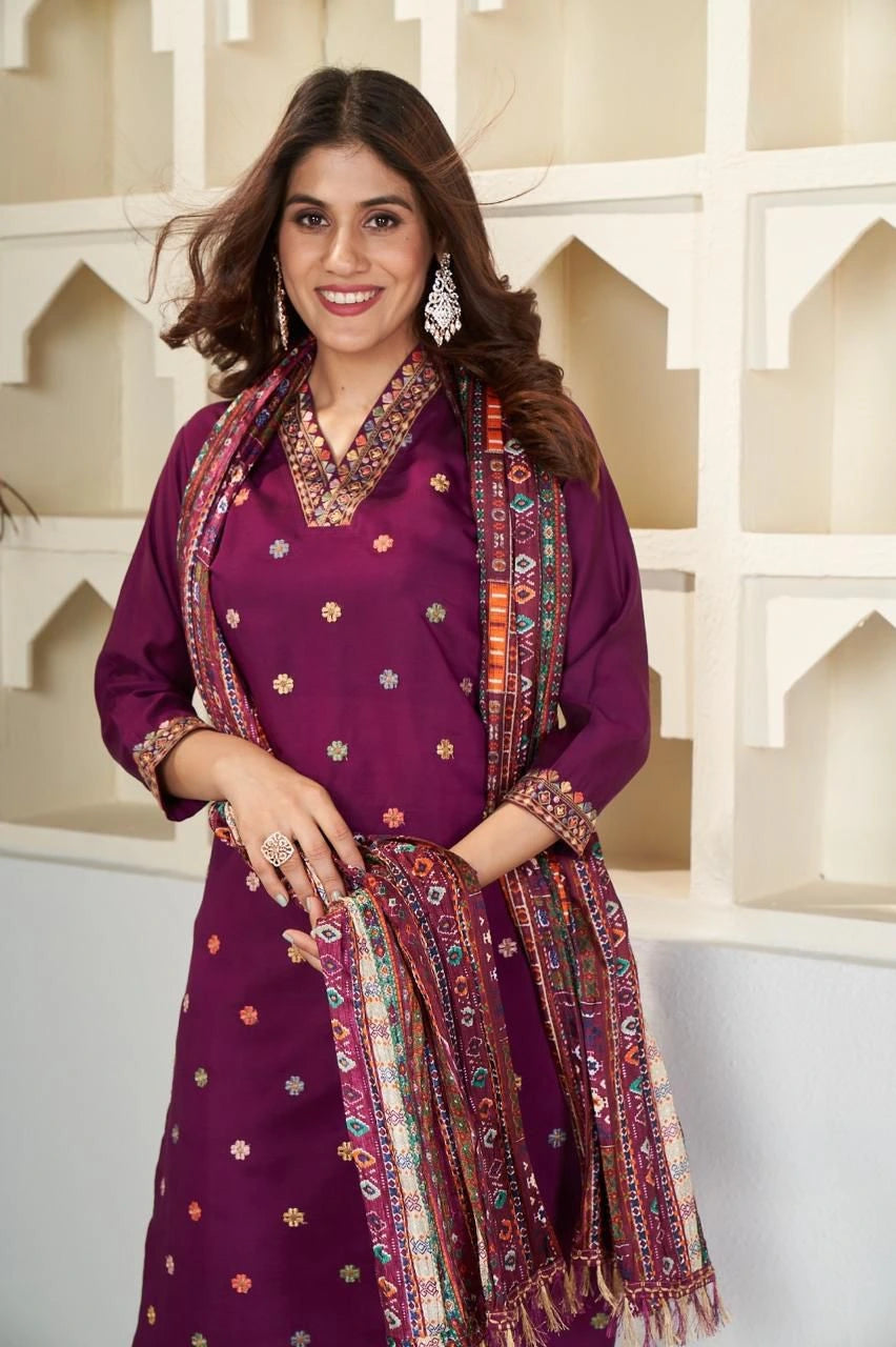 Woman wearing a traditional outfit with a purple top and patterned scarf against a decorative wall.