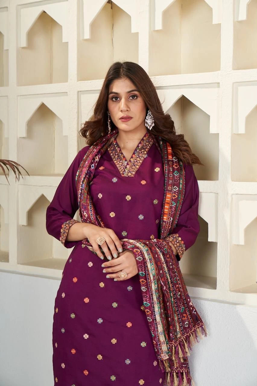 Woman in a purple traditional outfit with a patterned scarf against a decorative wall.