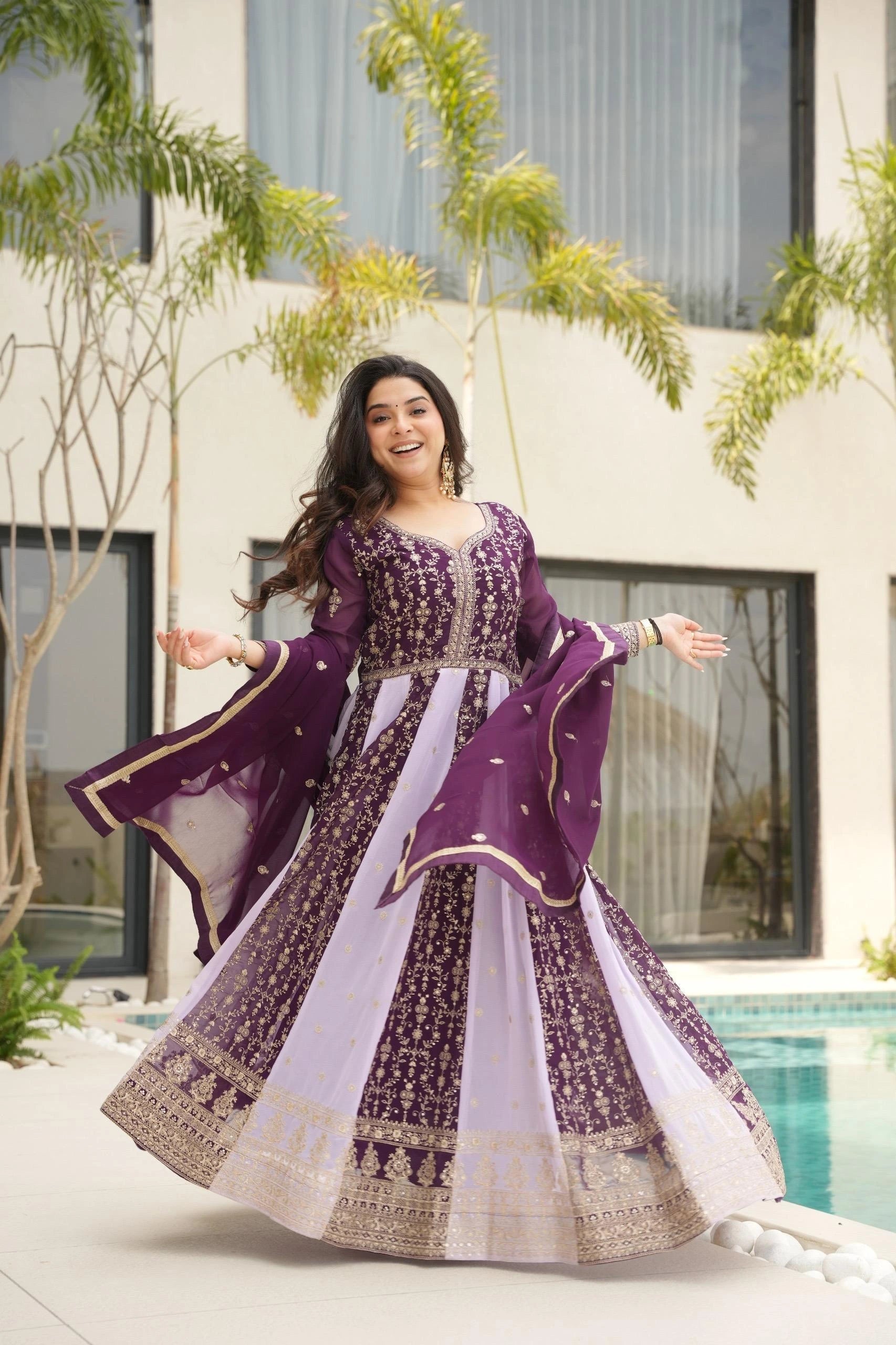 Woman in a purple and white traditional outfit standing by a pool with greenery in the background