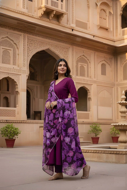 Woman in a purple outfit with a floral scarf standing in front of beige architectural background
