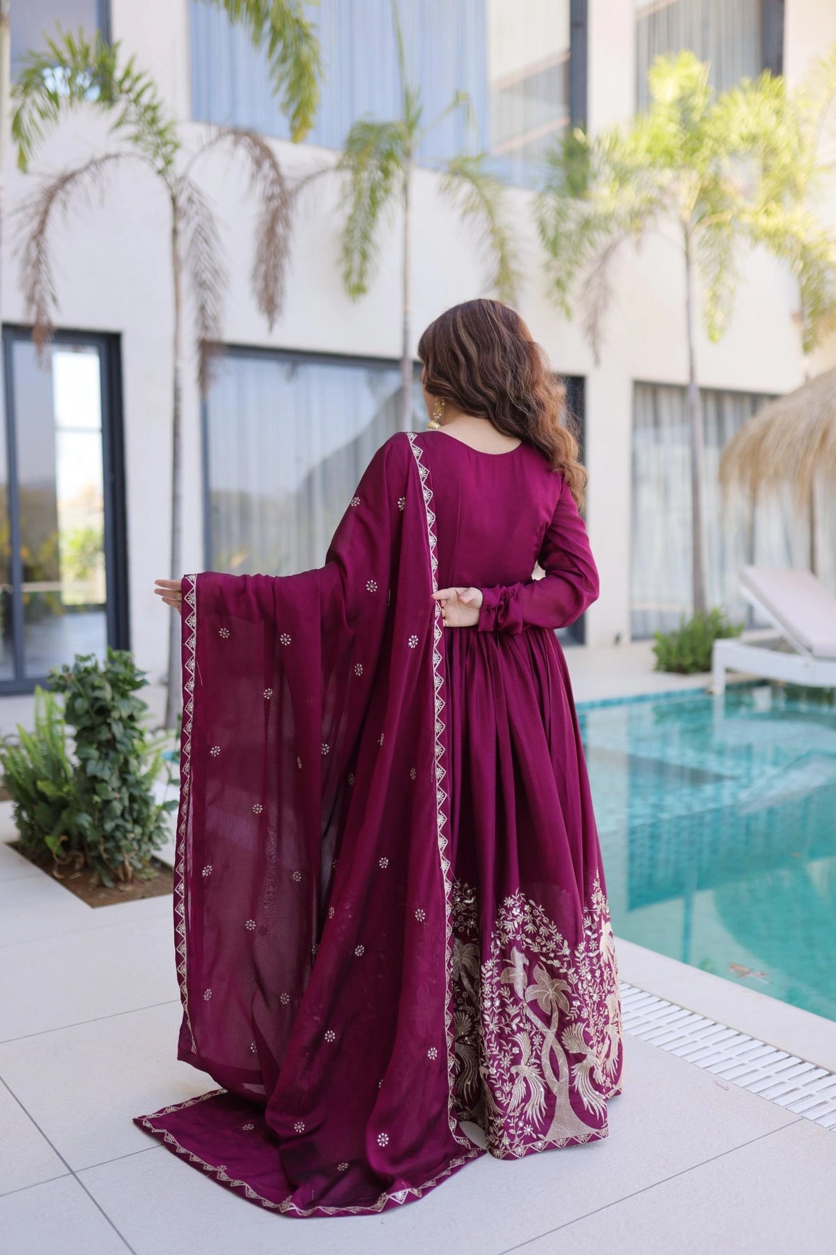 Woman in a burgundy traditional outfit by a poolside with modern building and greenery in the background