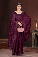 Woman in a purple saree with a matching blouse, holding a pink handbag against a dark background.