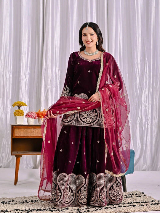 Woman in a traditional maroon and pink outfit with intricate designs, standing against a white curtain.