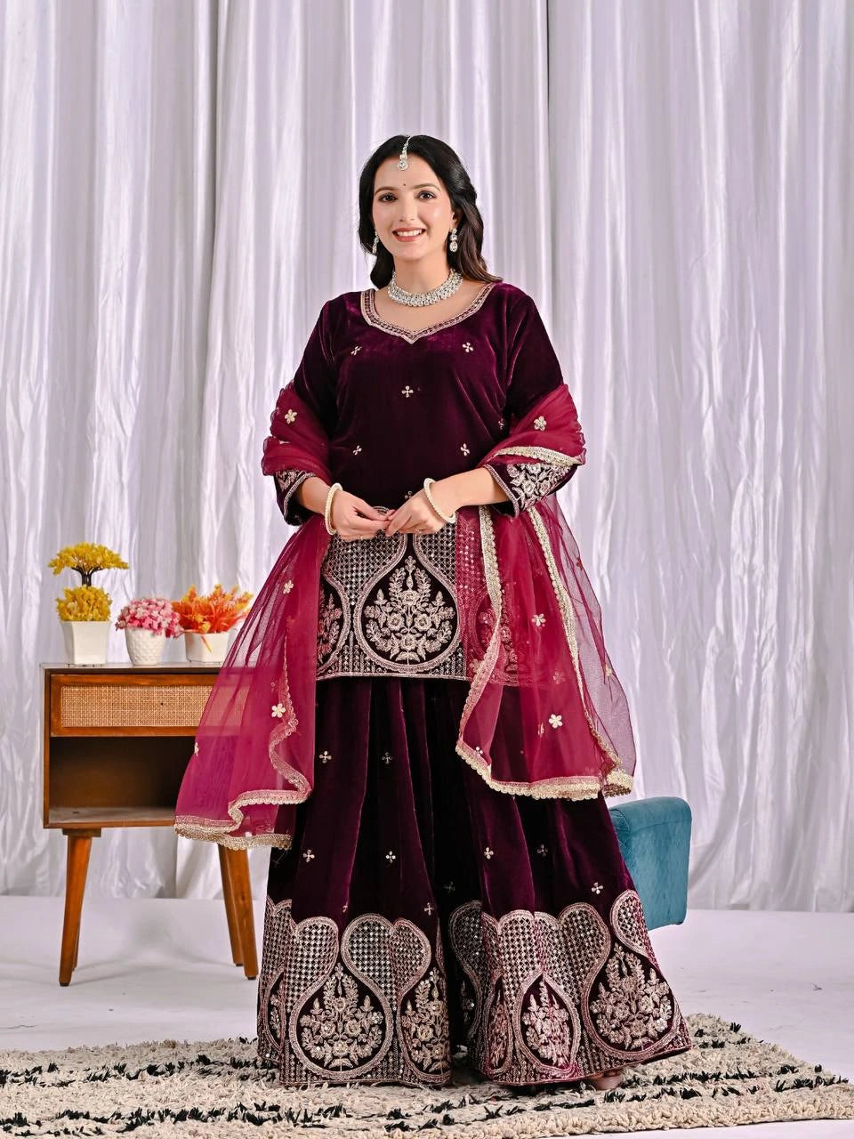 Woman in a maroon traditional outfit with intricate designs against a white curtain background