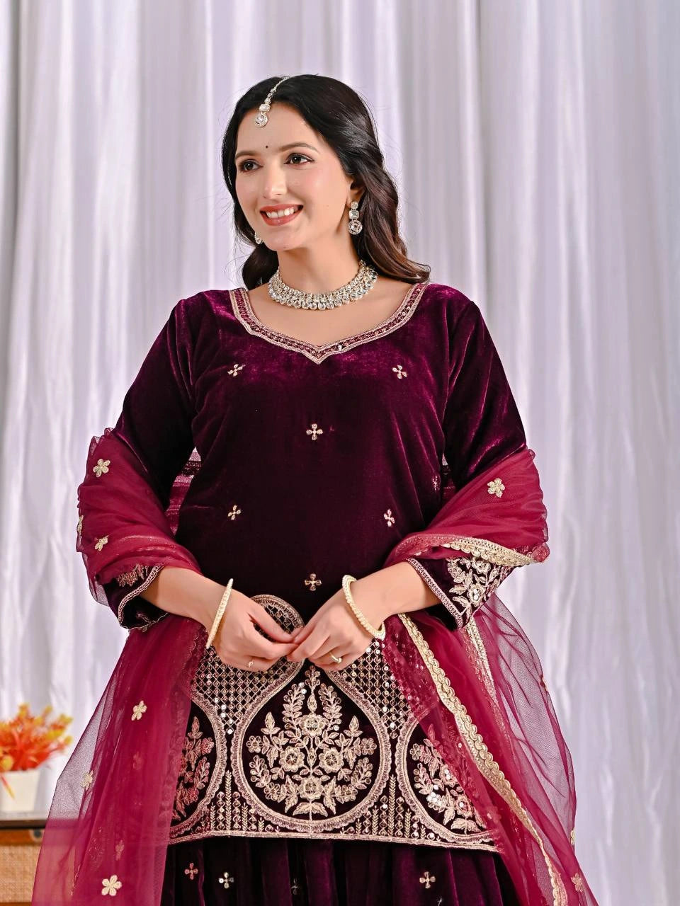 Woman wearing a maroon traditional outfit with intricate designs against a white curtain background