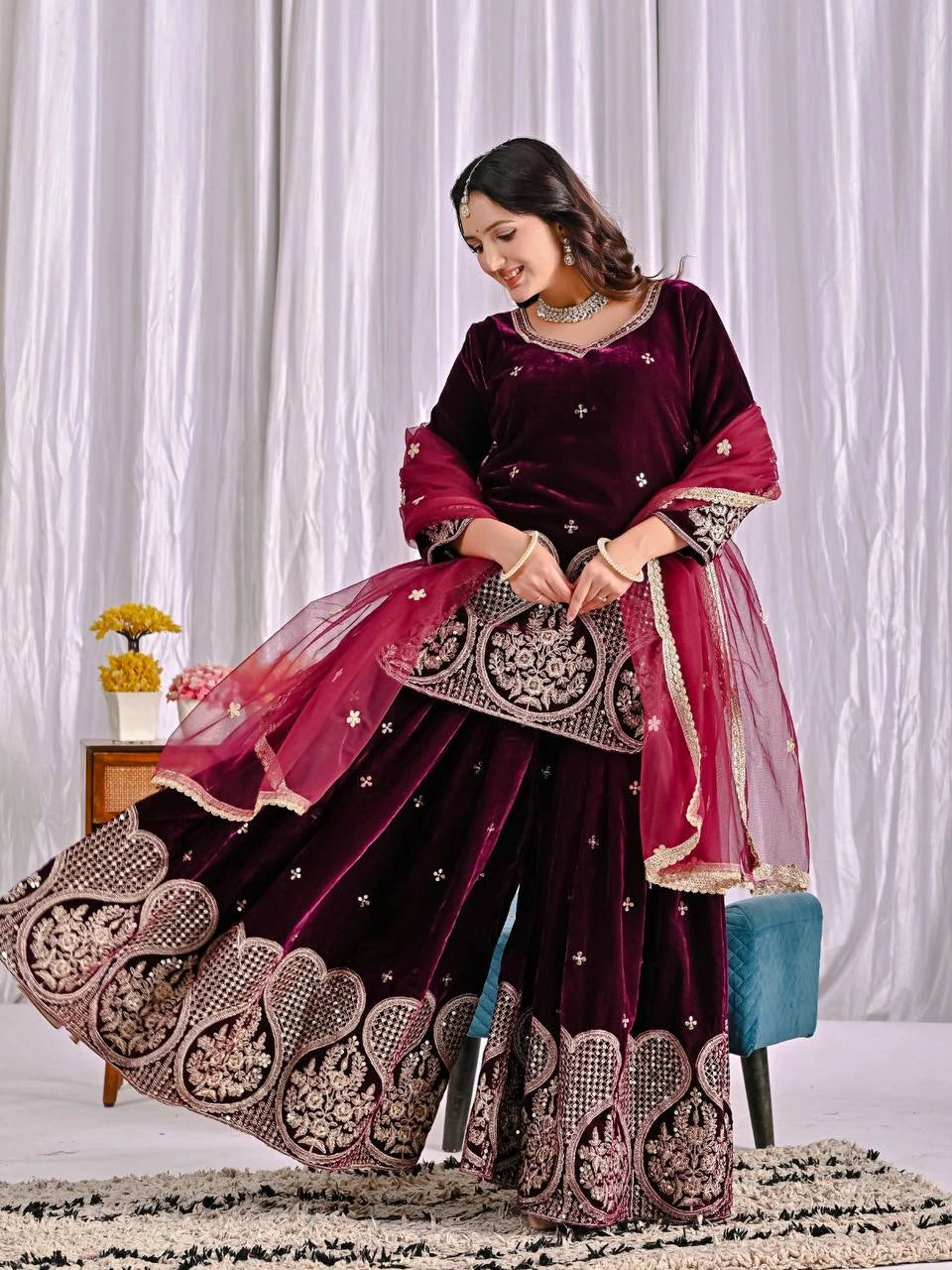 Woman in a dark purple and pink traditional outfit with intricate designs, sitting on a chair against a white curtain background.