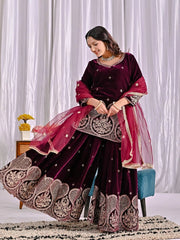 Woman in a dark purple and pink traditional outfit with intricate designs, sitting on a chair against a white curtain background.