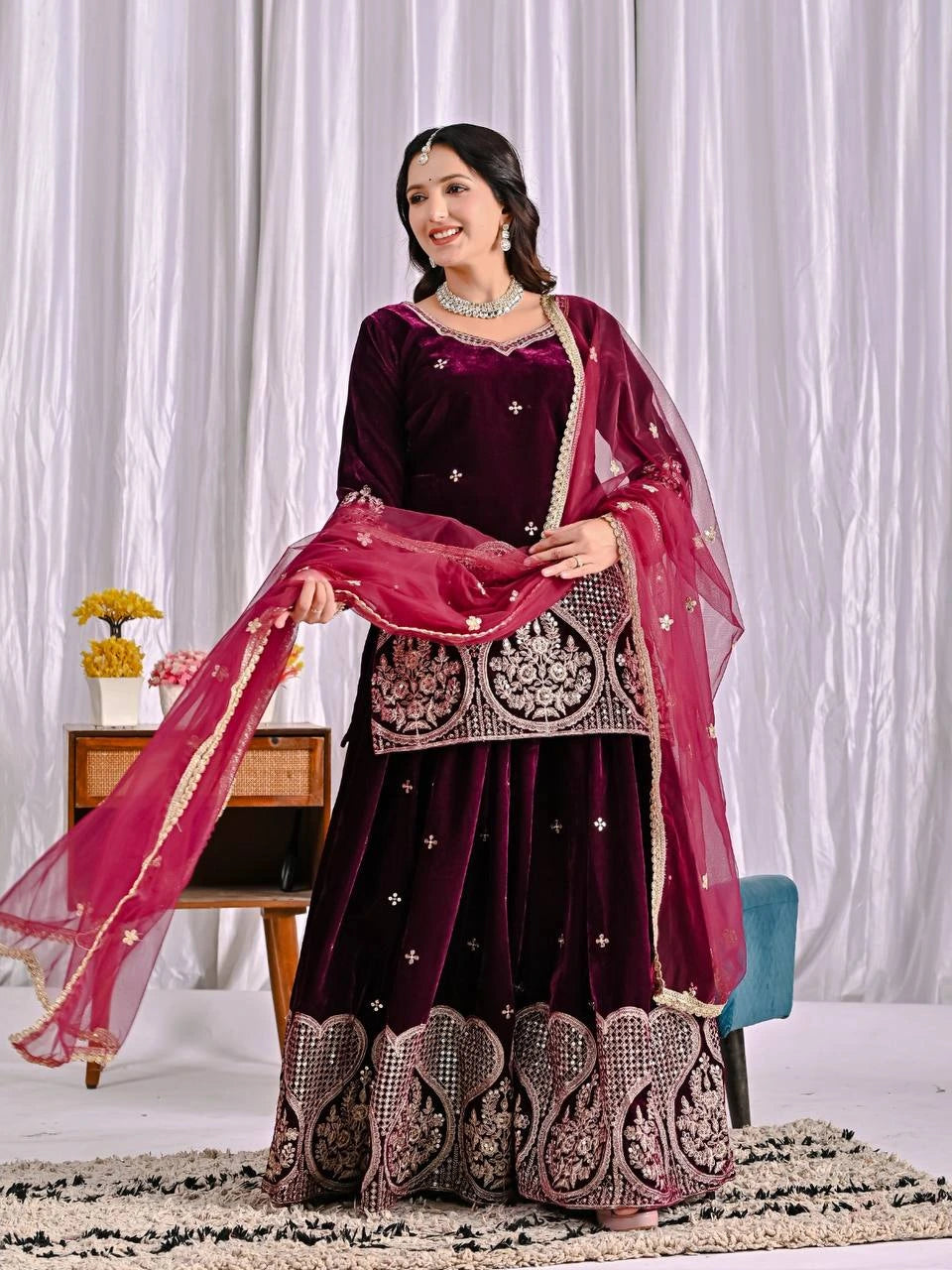 Woman in a dark purple traditional outfit with pink dupatta against a white curtain background
