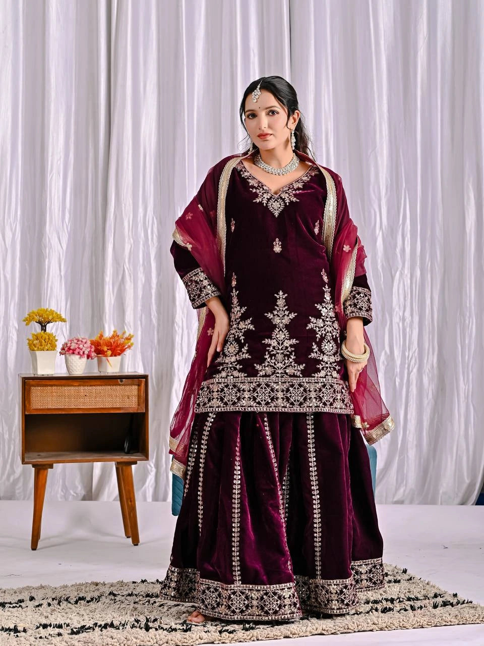 Woman in a maroon embroidered traditional outfit with a red dupatta against a white curtain background.