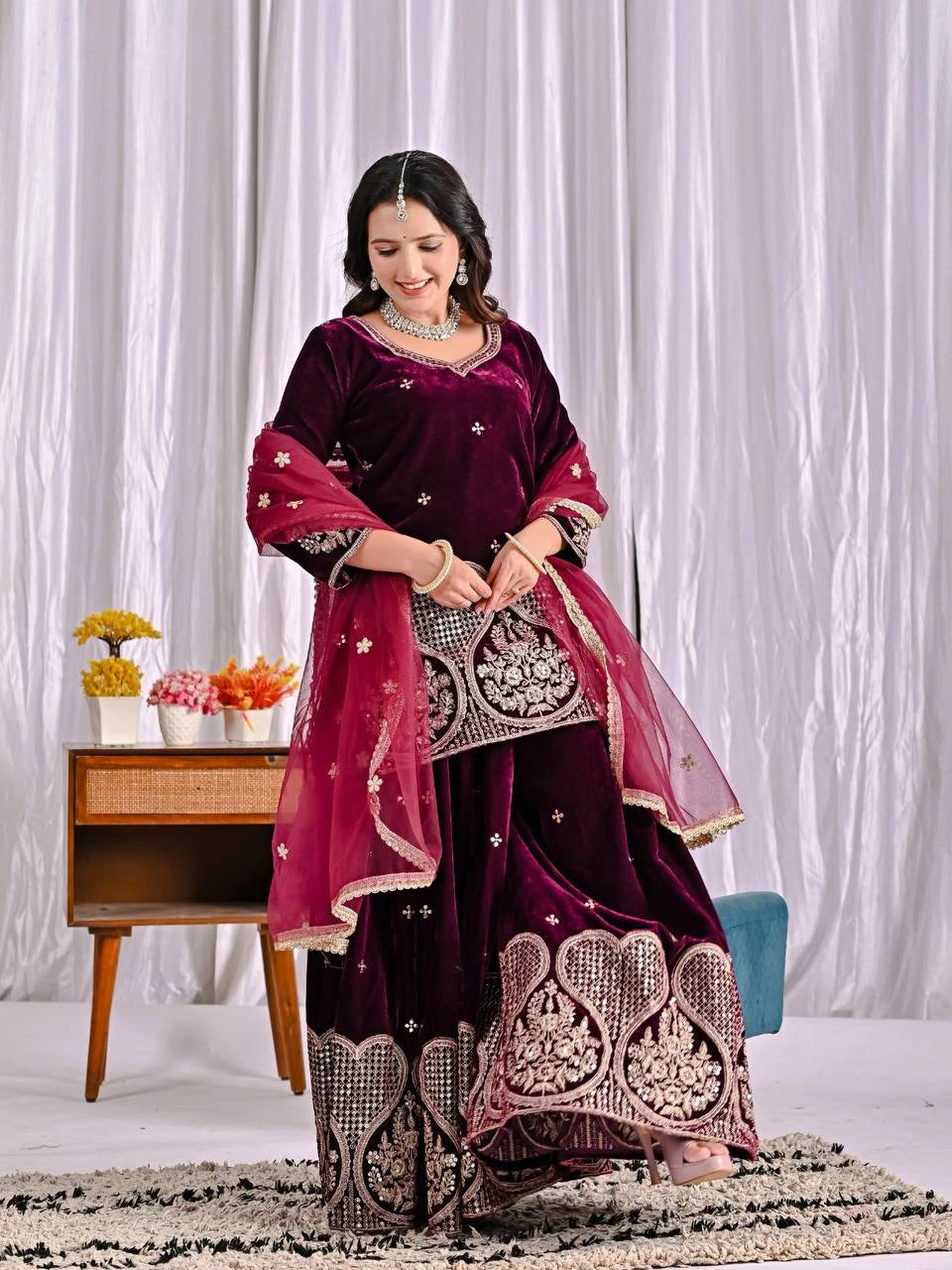 Woman in a maroon traditional outfit with intricate designs standing against a white curtain background.