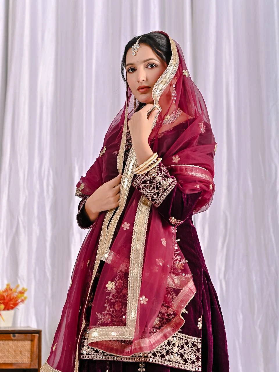 Woman wearing a maroon traditional outfit with a matching dupatta against a white curtain background