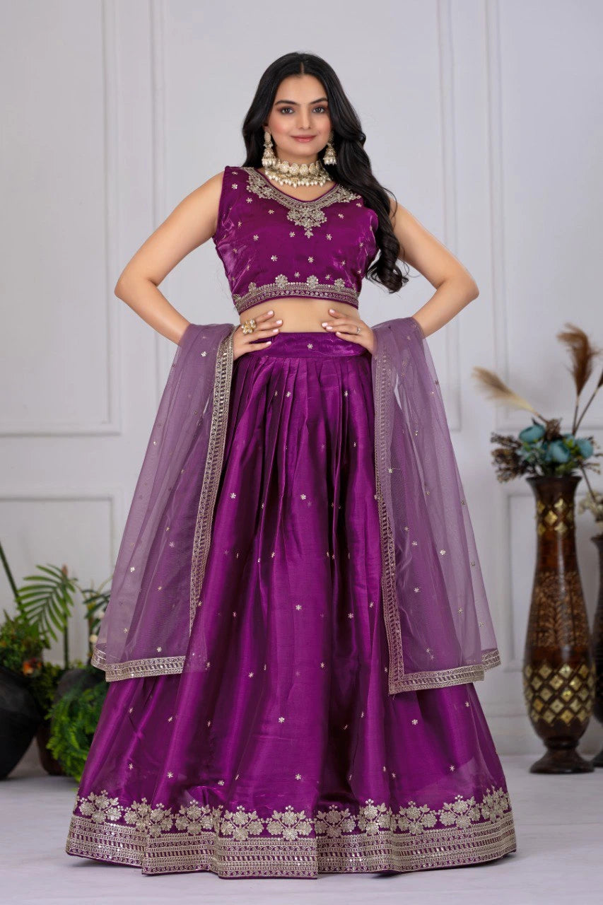 Woman wearing a purple traditional outfit with a matching dupatta against a white wall.