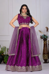 Woman wearing a purple traditional outfit with a matching dupatta against a white wall.