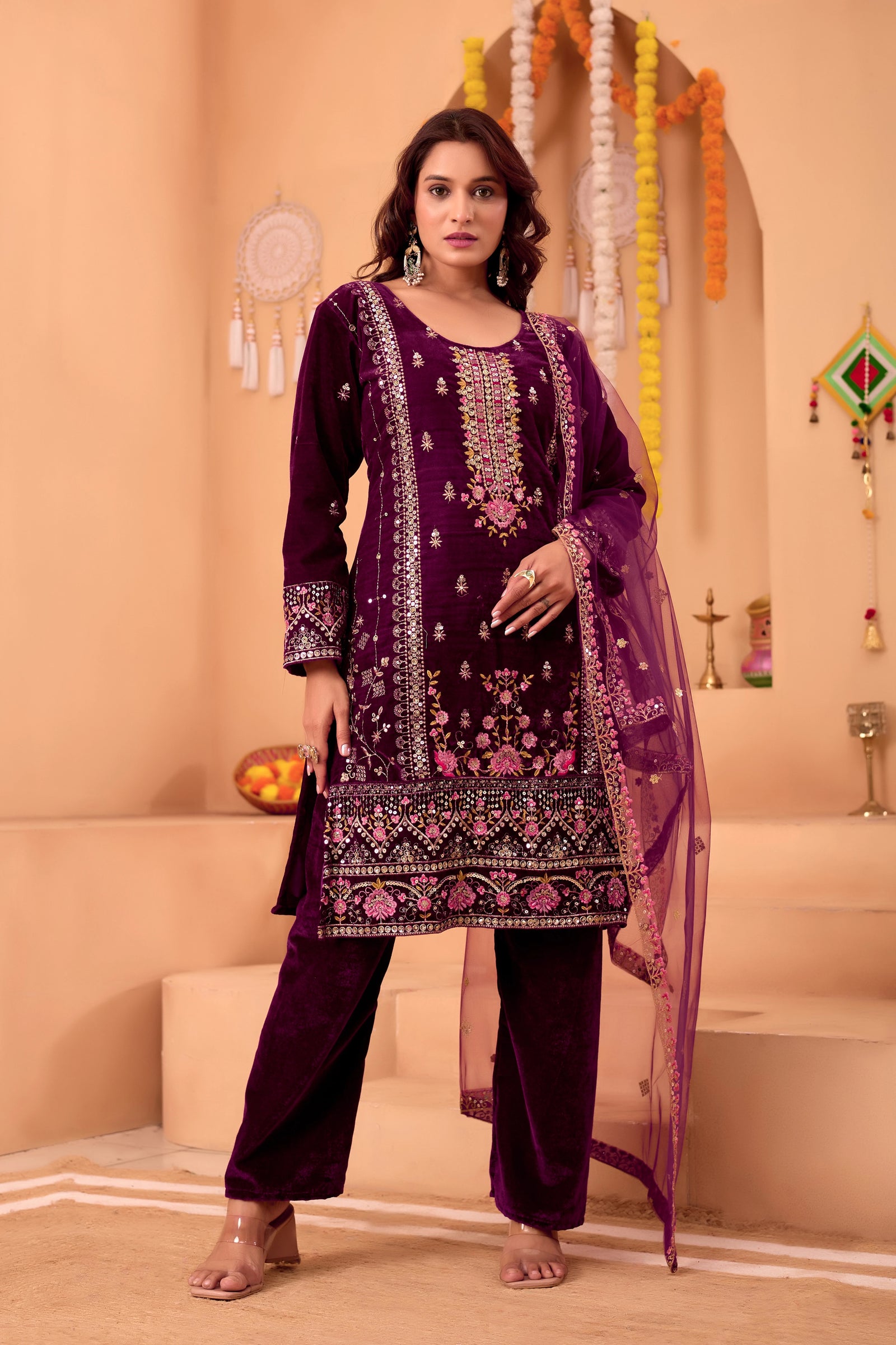 Woman wearing a purple embroidered traditional outfit with a pink dupatta in a decorated room.