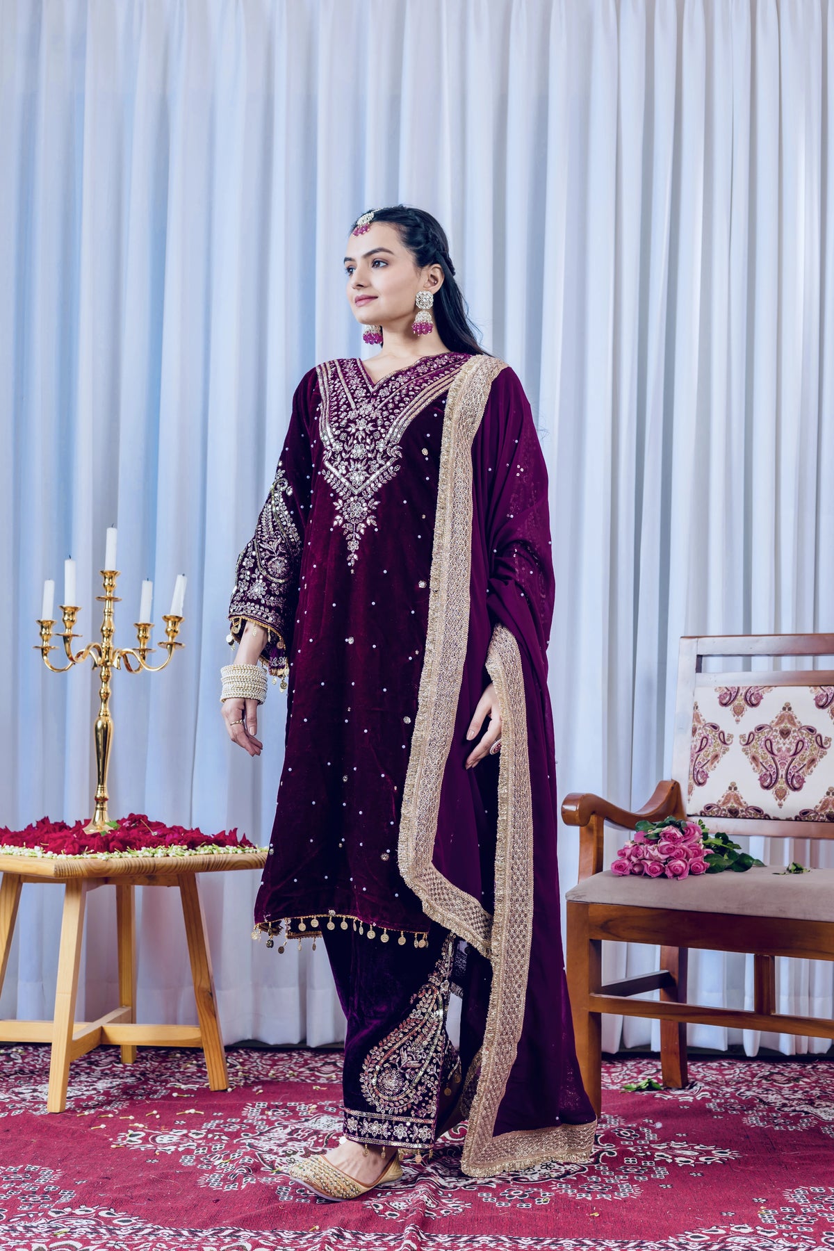 Woman in a dark purple embroidered traditional outfit standing in a decorated room with a candelabra and floral arrangements.