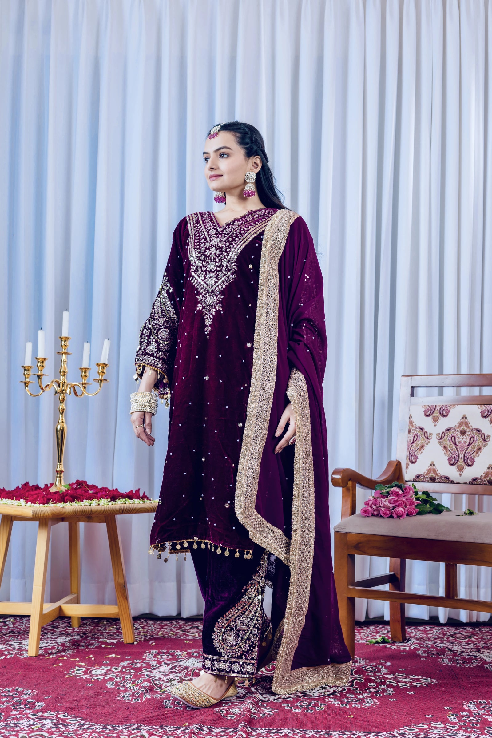 Woman in a dark purple embroidered traditional outfit standing in a decorated room with a candelabra and floral arrangements.