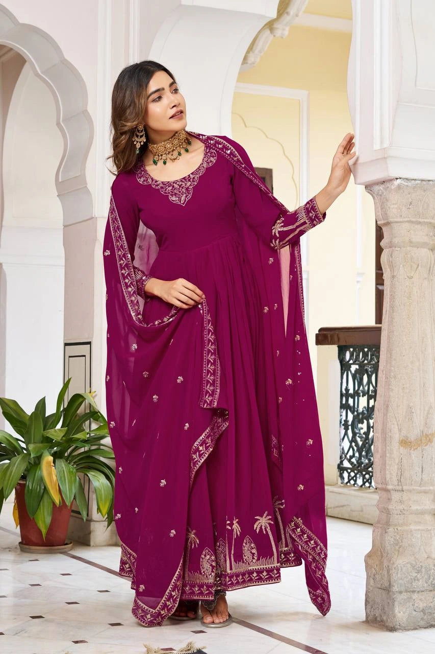 A full-length shot of a woman standing in a traditional ivory hallway, wearing a vibrant magenta Anarkali suit. The floor-length dress features a flared skirt with gold palm tree motifs embroidered along the hem and a matching sheer dupatta draped over her shoulders.