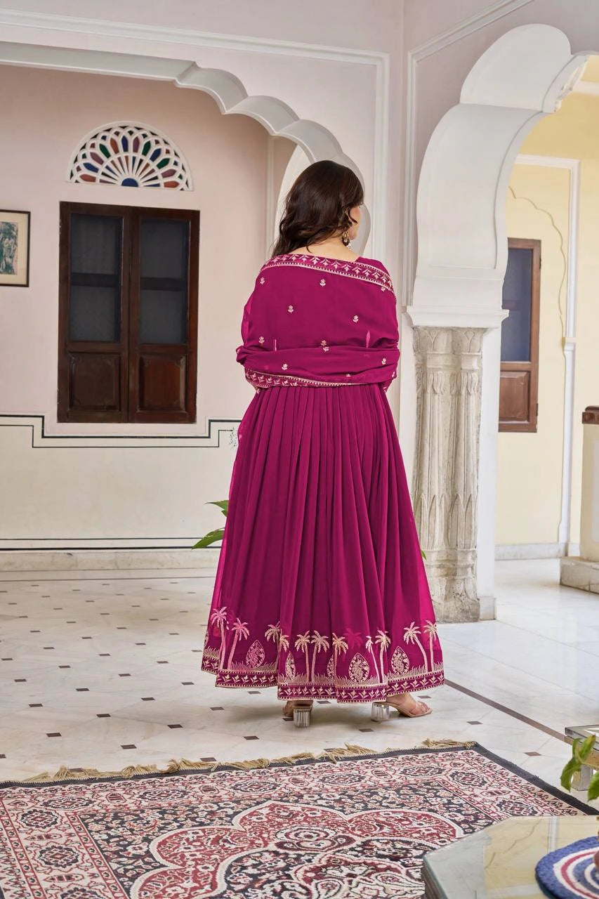 A full-length back view of the woman standing in a courtyard, highlighting the heavy pleating of the Anarkali that adds fullness to the skirt. The dupatta is wrapped around her back, showing the consistent gold floral buttis and border.