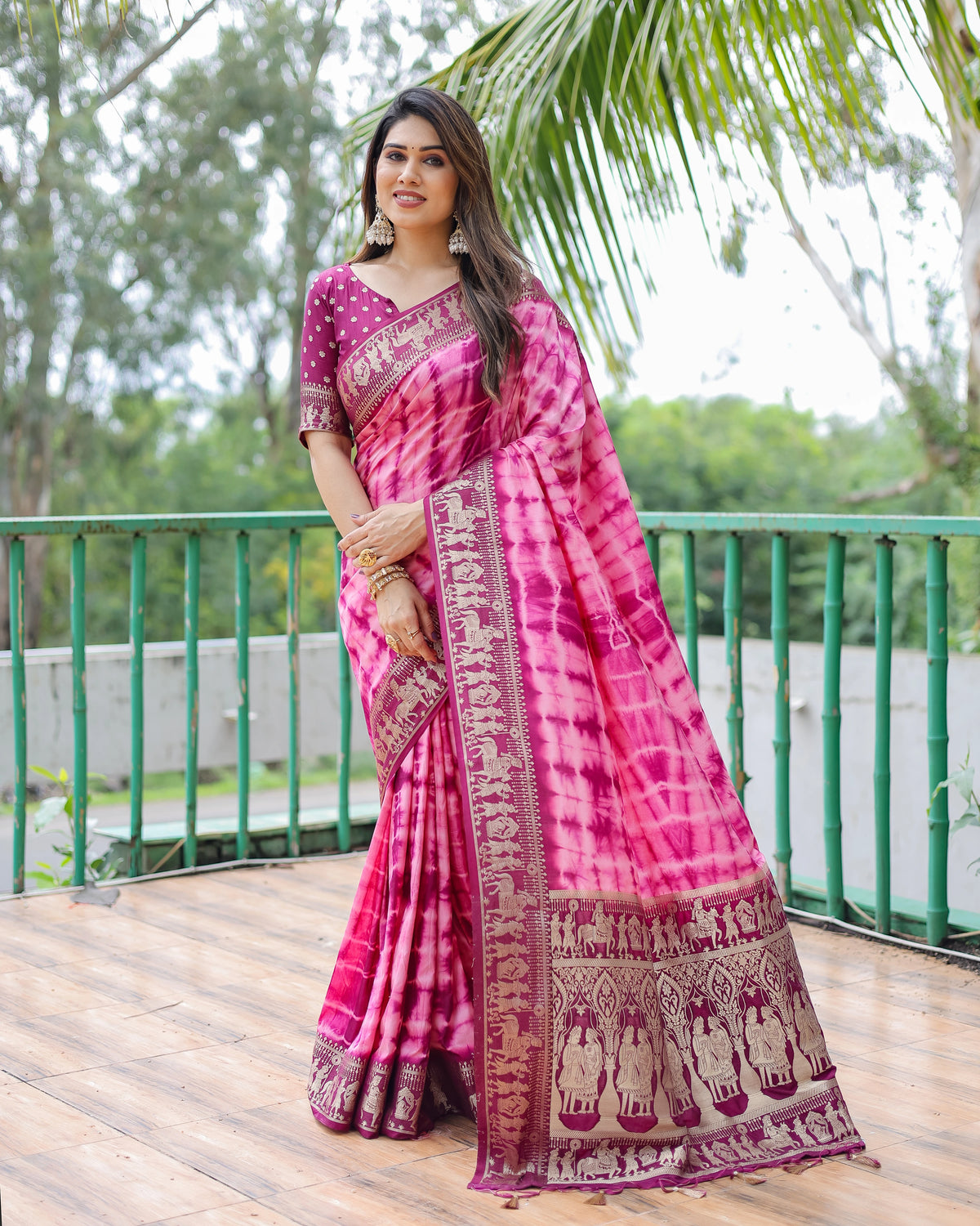 Woman wearing a pink saree with a traditional pattern on a wooden deck with greenery in the background