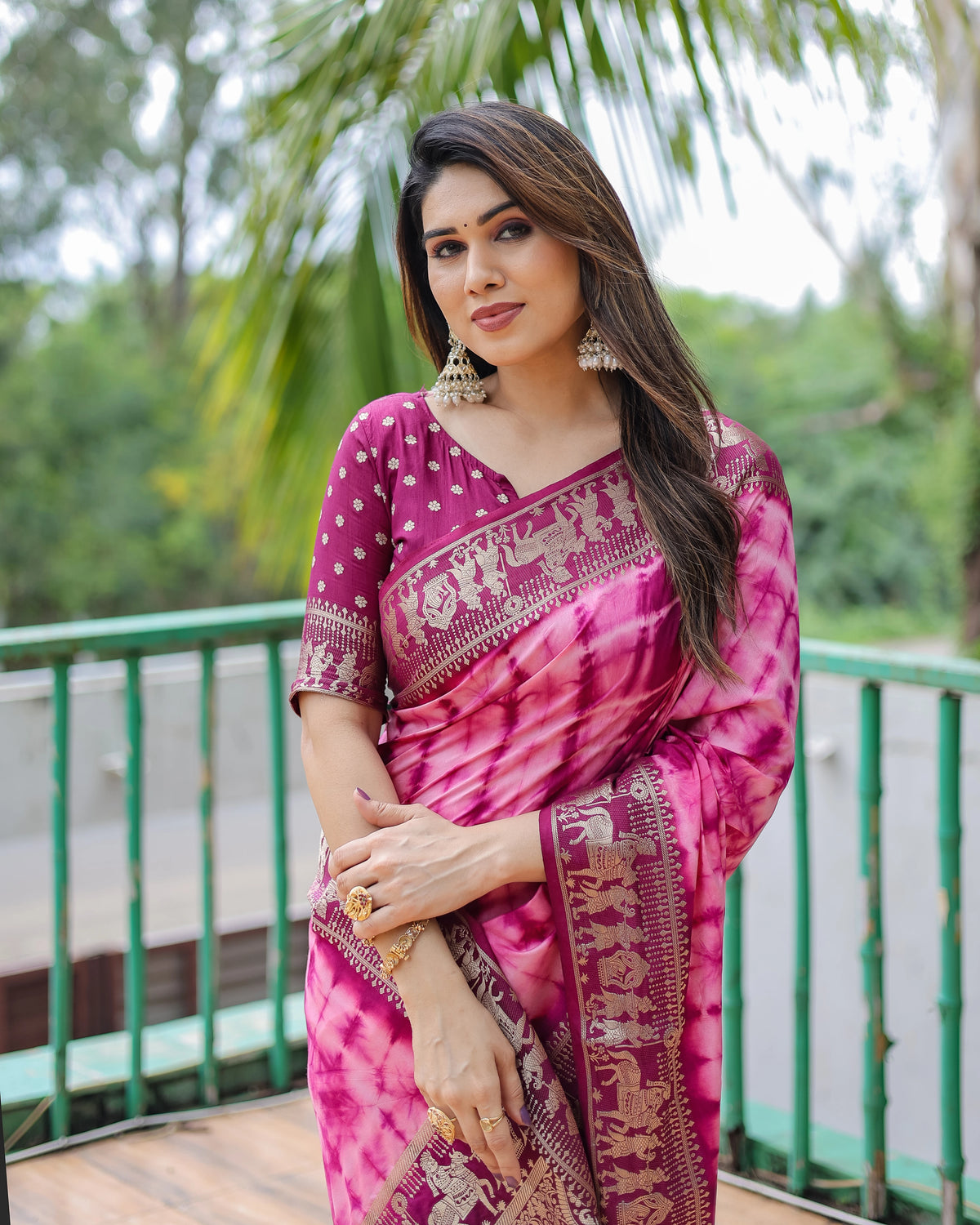 Woman wearing a pink saree with a traditional design, standing outdoors with greenery in the background.