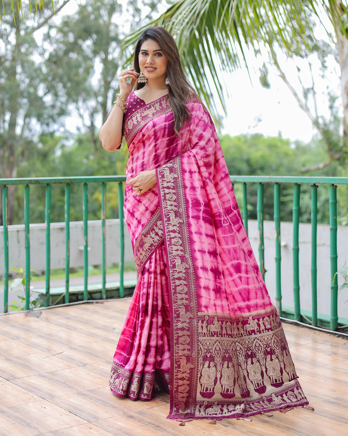 Woman in a pink saree with elephant patterns standing on a wooden deck with greenery in the background