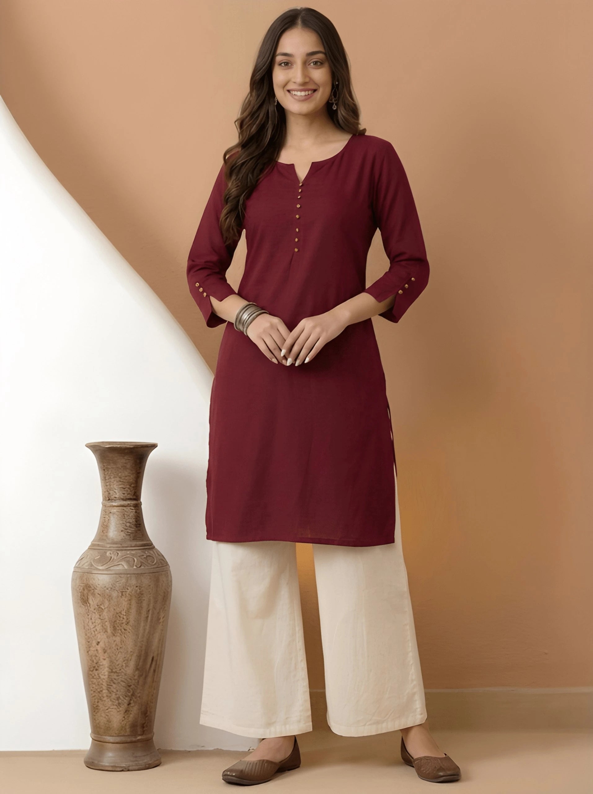 Woman wearing a maroon kurta with white pants standing against a gradient wall.